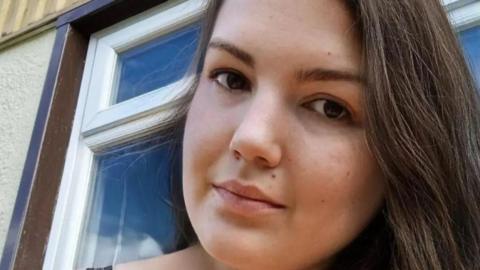 Sonia has long dark hair and brown eyes. She looks at the camera in a selfie.
