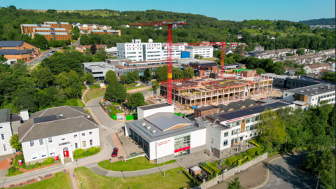 Campws prifysgol yn dangos adeiladau gwyn a gwaith adeiladu a dau graen yng nghanol y safle