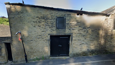 An old stone building which is blackened by soot and has several small windows which appear to be boarded up with black boards