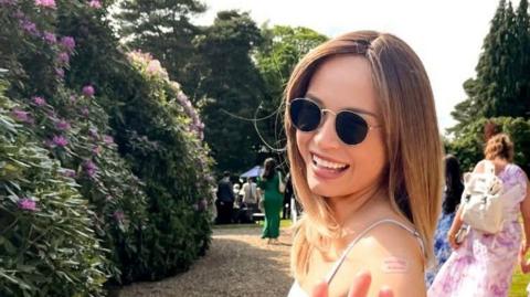 A woman in a garden wearing sunglasses, she is smiling at the camera.