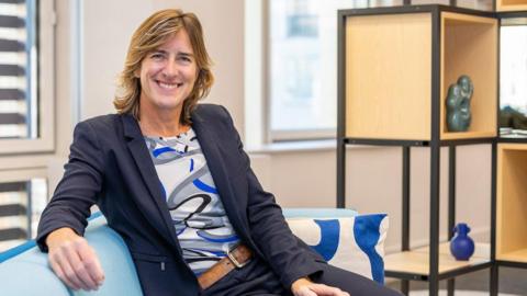 Dame Katherine Grainger sits in an office smiling at the camera. She is sat on a pale blue sofa, with her arm resting over the top of it and wears a navy blue suit, with a tan leather belt and a swirling patterned top in grey, blue and black and has dark blonde mid length hair. In the background is a wood and balck metal shelving unit which has a number of different ornaments in the shelves, they are slightly out of focus behind her.