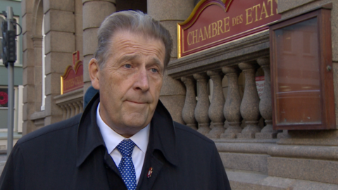 A man looking at the camera wearing a black coat, white shirt and blue spotty tie standing in front of a government building.
