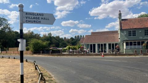 A village road sign points to the village of Ringland on the corner of a road, with a pub in view behind it, on a sunny day.