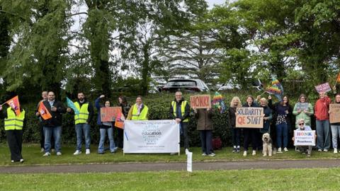 A group of people wearing hats and coats stand on the grass outside a school holding signs with slogans such as "Save our teachers" and "Stop job cuts". The picture is taken from across the road. There is also a dog in the line. It is an overcast day.