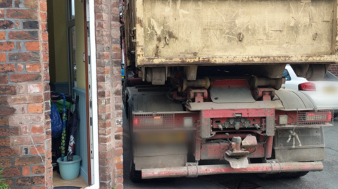 A lorry on the pavement and very close to the wall of a house