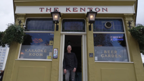 The pub is a yellowish/green colour with old fashioned signage that reads The Kenton and lamp lights. Signage in the windows reads "karaoke room" and "beer garden". A man with grey hair and wearing a black shirt and grey jeans stands in the doorway. The camera is pointing at an angle upwards from the ground. 