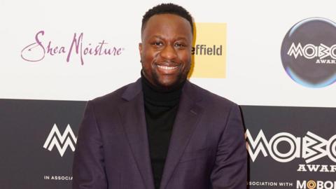 A man in a black suit jacket and black polo-neck stands in front of a black hoarding for the Mobo Awards. He has black cropped hair shaved around the sides. He is smiling.