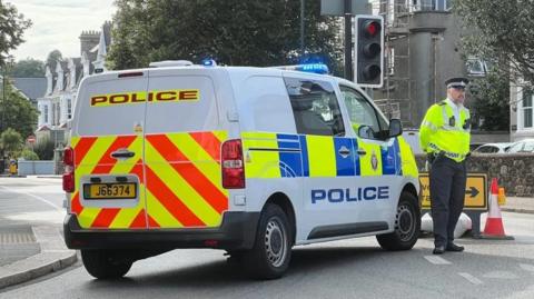 A policeman in a high visibility jacket stands by a police van  at a road junction.