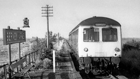 A black and white image of an old train on a railway line running through a field. A sign on the front of the train reads 'Yarmouth'. To the left of the railway line there is a bench with a sign above it that reads 'Berney Arms'.