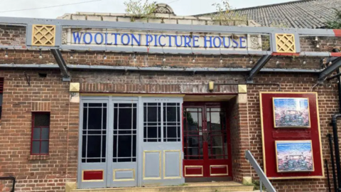 Outside of the Woolton Picture House. It's an old bricked building with painted red doors. A carved text reading the name of the cinema is above the doors.
