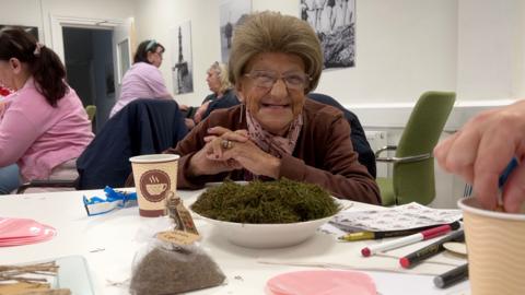 An elderly woman smiles while sat at a craft table with a bowl of turf from the Sunderland AFC pitch on it.