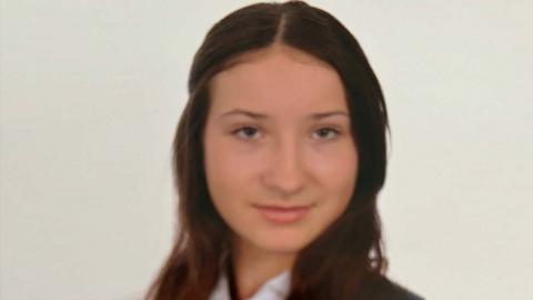 A teenage girl with long brown hair looks at the camera. She is wearing a white shirt, a navy and white striped tie and a navy blazer.