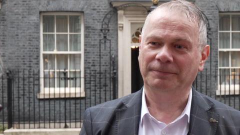 Corin Dalby, with receding grey hair and wearing a white shirt and grey suit with a lighter grey pattern, stands in the rain across the road from 10 Downing Street.