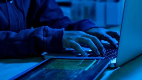 An anonymous pair of male hands hover over a laptop keyboard. A tablet with green writing is to on-side of the laptop.