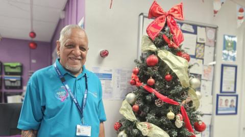 An older gentleman, in his 70s, stands smiling in front of a beautifully adorned Christmas tree and a notice board pinned with cards. He is South Asian. He wears a blue t shirt and a lanyard, both of which identify him as an NHS volunteer. The space around him is festooned with Christmas decorations. 