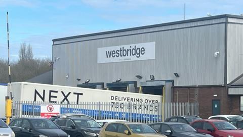 A factory building with a sign saying 'westbridge'. A next lorry is parked outside. Multiple cars are parked in a fenced off car park next to it. 