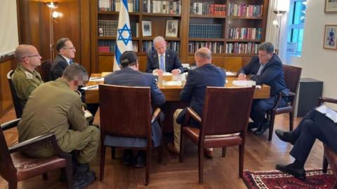 PM Benjamin Netanyahu pictured during the phone call with president Biden. He sits at a wooden table surrounded by seven aides, with an Israeli flag visible behind him.