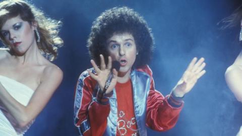 A man, Leo Sayer, wearing a blue and red sparkly top while singing into a microphone.