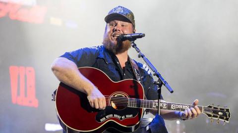 Luke Combs performs during 2025 Austin City Limits Music Festival at Zilker Park. He has a brown beard is wearing a camo baseball cap, wearing a black short sleeve shirt and playing a guitar singing into a microphone.
