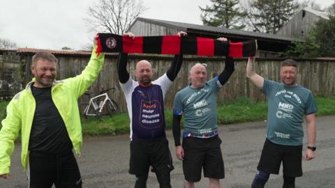 Four men holding up a Truro United scarf as they arrive in Truro, Cornwall. The scarf is red and black. They are all smiling at the camera. 