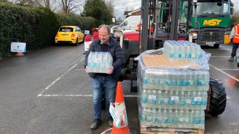 A man carrying a large pack of water bottles. There is an even larger stack of water bottles next to him on some heavy machinery, as well as vehicles in the background in what appears to be a car park. 