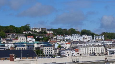 A view of St Peter Port with an array of tall, narrow three and four storey buildings, and a line of trees in the background, on a bright day.