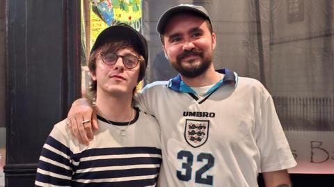 George Wright, wearing a Euro 96 England shirt with 32 on the front, puts his arm around his brother, Laurie, wearing a stripey long-sleeve t-shirt while standing outside a pub in central London. 