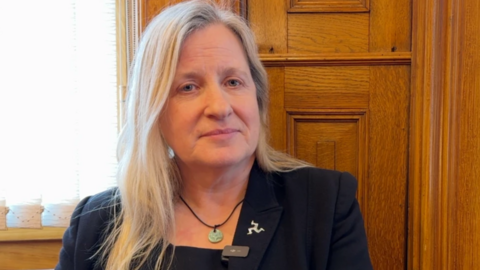 Michelle Haywood has long, straight blonde hair. She is wearing a dark suit and sitting in front of a wooden interior wall with a window to the left.
