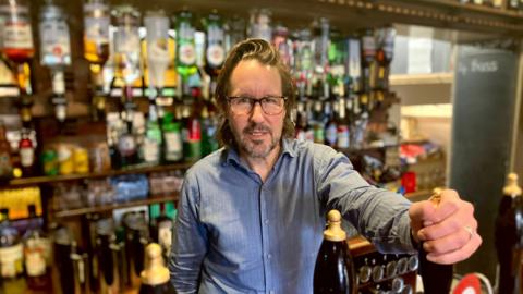 A man with long brown hair, glasses and dark facial hair is standing behind a bar holding onto a beer tap handle. He is wearing a blue shirt and there are bottles on the wall in the background
