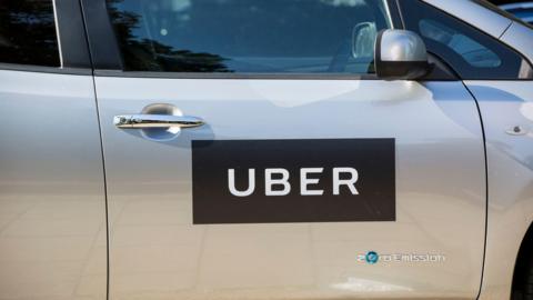 A Uber car photographed from the side. It is a light grey car with Uber's logo on the driver's door and a 'Zero Emission' writing