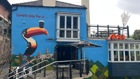 The Boat Club mural shows a large toucan balancing a pint on its beak next to the venue's entrance door. Durham Cathedral and the River Wear are pictured in the background. Most of the wall is blue. "Lovely day for a..." is written above the toucan. The building has a glassed terrace on its lower floor and an open one at the top. There is a bright orange Aperol sign on the roof of the indoor terrace. 