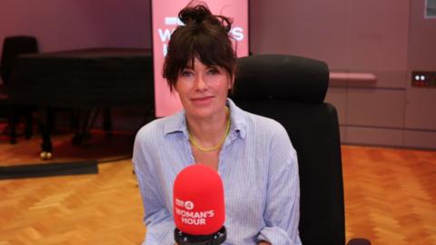 A woman in a brown-haired bun and linen blue shirt sits in front of a microphone.