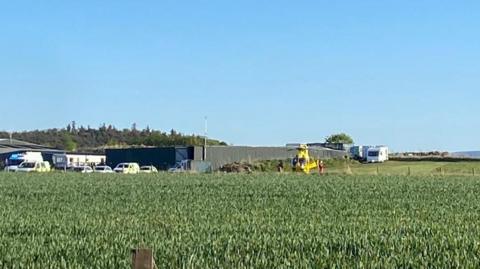 Several emergency service vehicles including an ambulance helicopter in the middle of a field. Campervans can be seen in the background