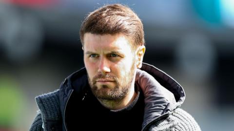 Brighton boss Fabian Hurzeler, in a hooded jacket and with dark brown hair and beard, looks on before a Premier League match at Burnley