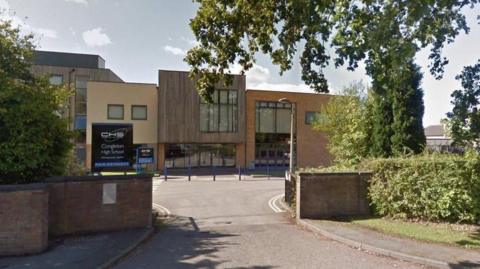 The front of a school building with a driveway leading up to it. There are blue bollards directly infront of a brown reception area