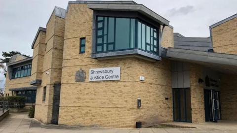The side of a large brown brick building with dark windows and a sign which reads Shrewsbury Justice Centre