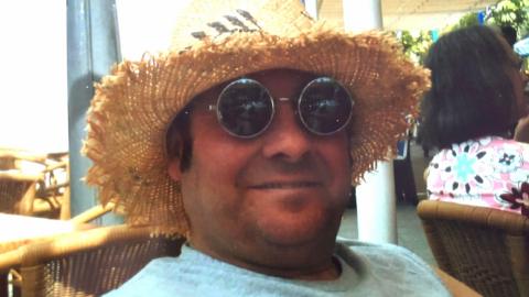 Robert Newcombe smiles at the camera and is wearing a straw hat and a pair of sunglasses. He has a grey vest on. 