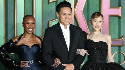 Cynthia Erivo, John M. Chu and Ariana Grande stand in front of a Wicked logo at the film's European premiere in London.