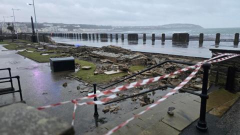 Picture of the damaged Sunken Gardens. Red and white tape marking off the sight can be seen whilst a bricked wall lies on the ground after being blown over. Picture taken on a grey, wet day.