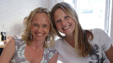 Two women sit together and smile at the camera. Their heads are leaning together and they are smiling. They are in a restaurant and behind them is a table with a wine bottle and wine glass on it.