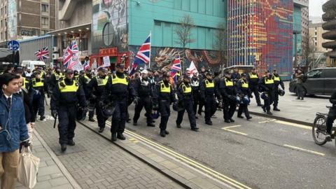 A small group of people march with Union flags and england flags while being escorted by a large group of police
