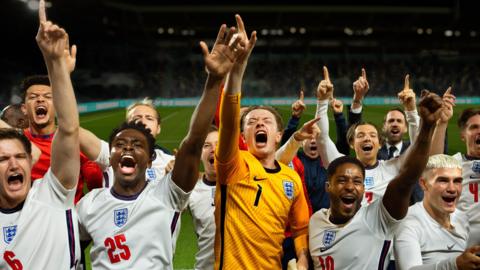 In a scene from Dear England, a team of footballers wearing the England men's team white and red kit celebrate on a football pitch, their goalkeeper, wearing yellow stands in the middle.