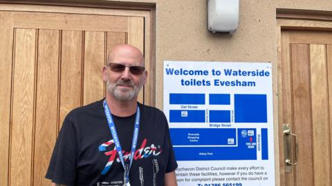Phil Childs wearing a black t-shirt and a blue lanyard while stood in front of a black of toilets with light brown wooden doors. There is also a sign that reads: "Welcome to Waterside toilets Evesham. Phil has a bald head and a grey beard. He is also wearing sunglasses. 