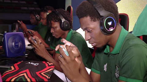 Gamers wearing headphones and green shirts huddle over handheld screens