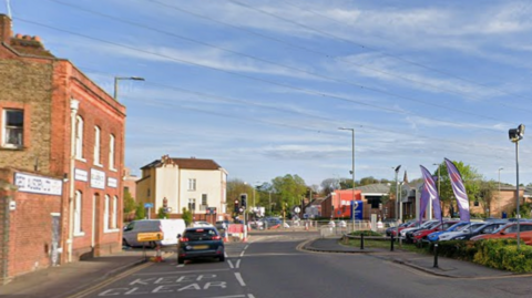 Part of Lower High Street in Watford. There are cars approaching a junction. On one side is a brick building and cars at a dealership parked on the right. 