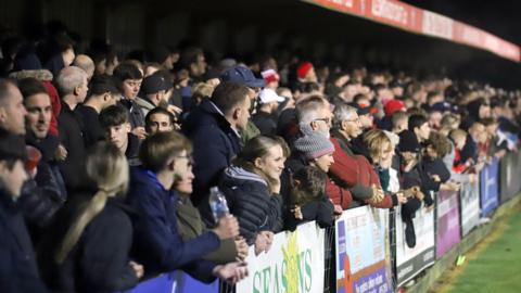 Hundreds of Kettering Town fans packed into a stand at their ground. They are looking away from the camera and at the action on the pitch. The fans in the front row are leaning over the hoardings.