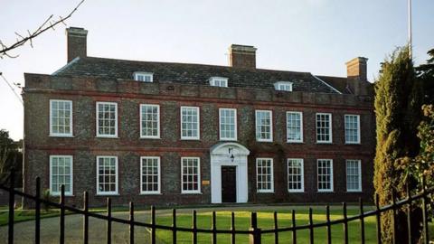 Sussex Police's headquarters in Lewes. A two-floor brown brick building with three chimney stacks on the top and a white entrance over a black door. The photo is taken over black gates and there are trees and grass in the foreground