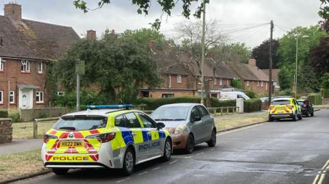 Two police cars are parked in Queen Mary Avenue, a road with semi-detached, two-storey, brick houses with steeply pitched roofs. 