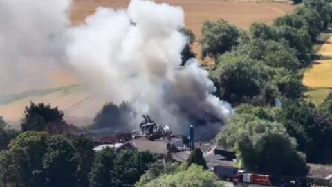 Aerial view of a large fire at a scrapyard. There is thick smoke billowing up from the blaze, and the area is surrounded by trees.