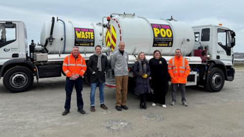 Six people - two in high visibility jackets - stand in a car park in front of sewage lorries with Let's Talk Poo and No Time to Waste banners on the sides.